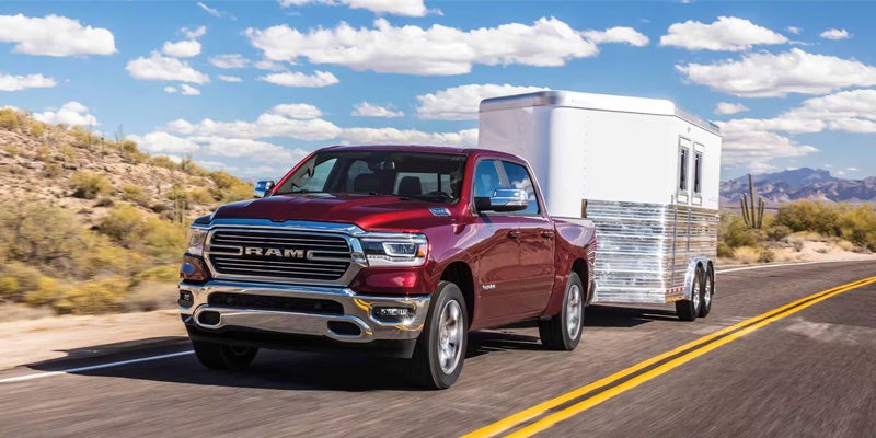 A red Ram pickup truck pulls a silver and white horse trailer down a desert highway on a sunny day.