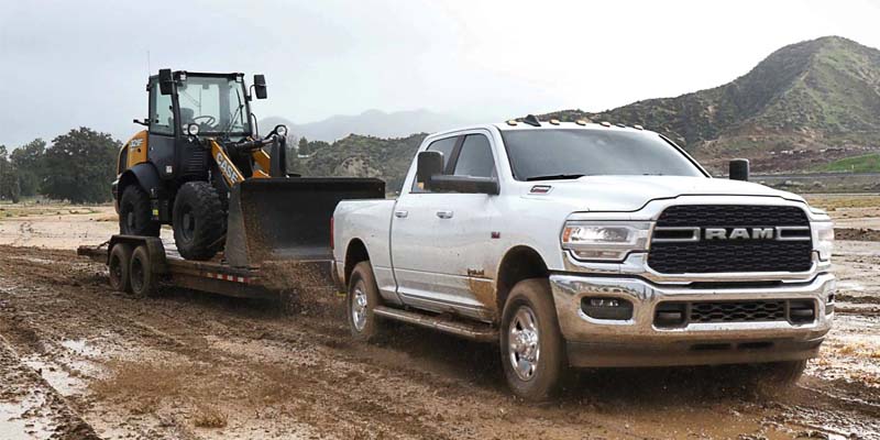 A white Ram truck pulling a trailer with a yellow Case front-end loader on a muddy dirt road.