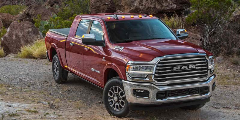 A maroon Ram 2500 heavy-duty pickup truck with a black bed cover, parked on a dirt path in a rocky, desert landscape.