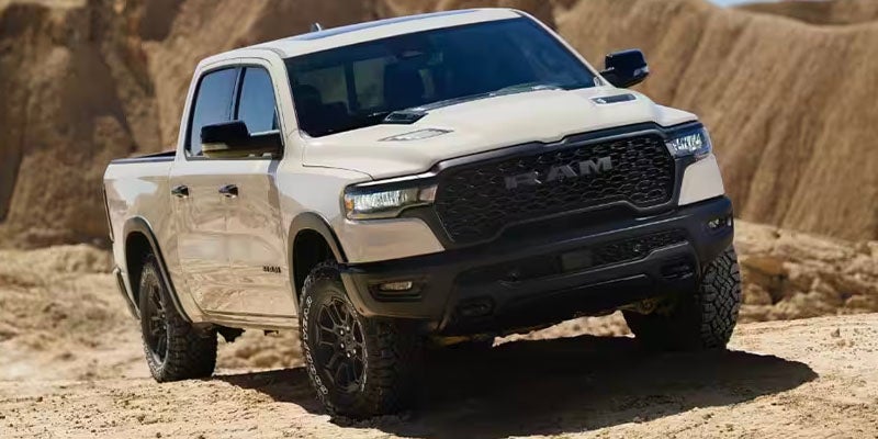 Off-white Ram TRX pickup truck on a sandy dune, angled left with rugged terrain in the background.