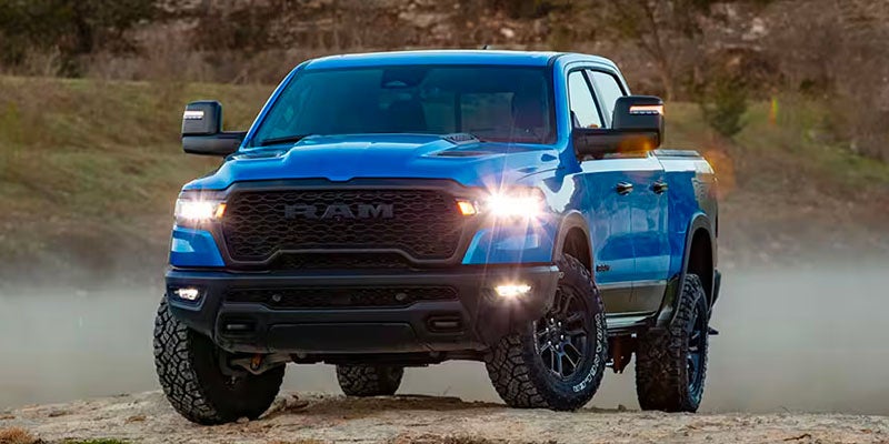 Bright blue Ram pickup truck with headlights on, parked on a dirt path with dust behind it.